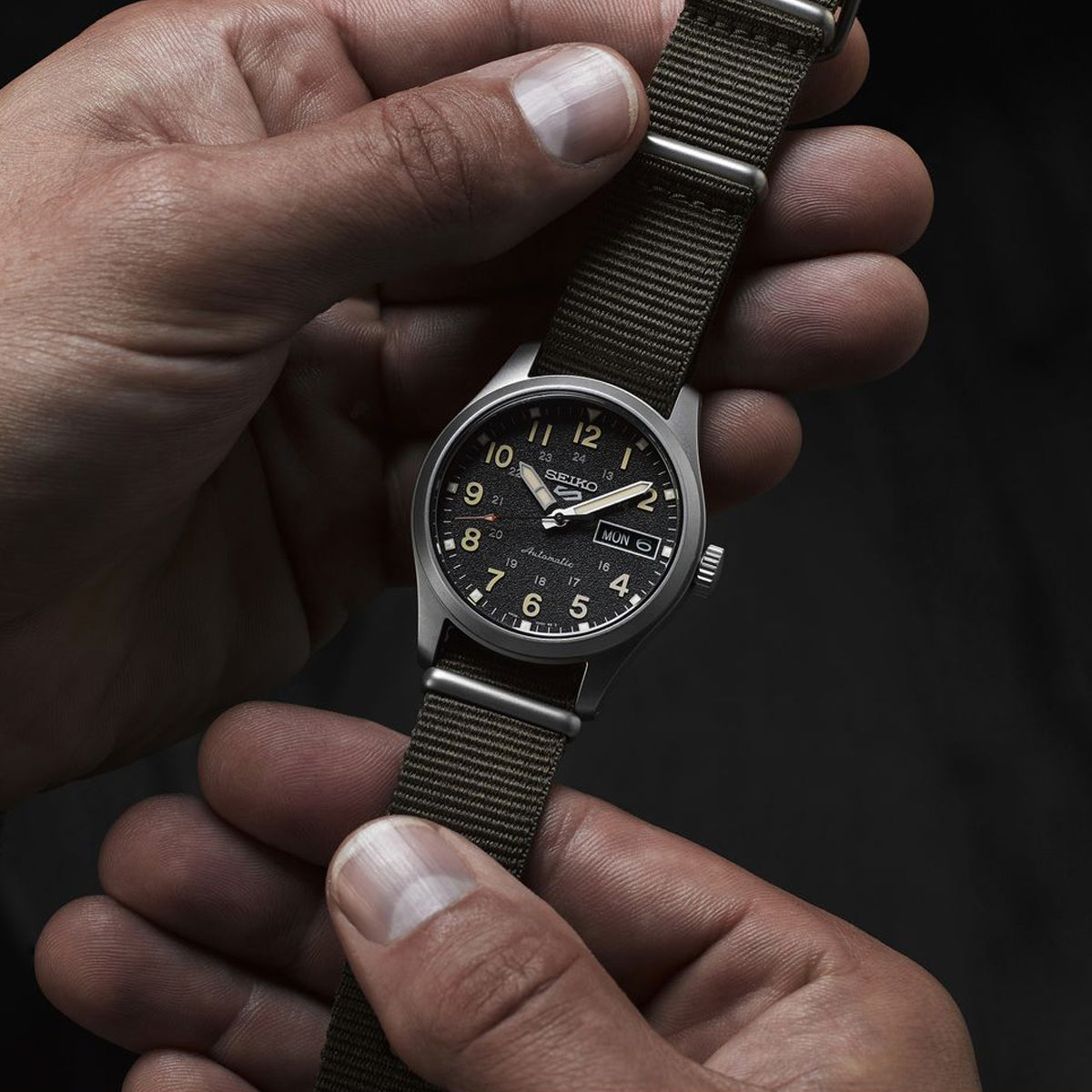 SEIKO MOD. SRPJ85K1 wristwatch with fabric strap, stainless steel case, mineral dial, showing day and date functions, held in hands.