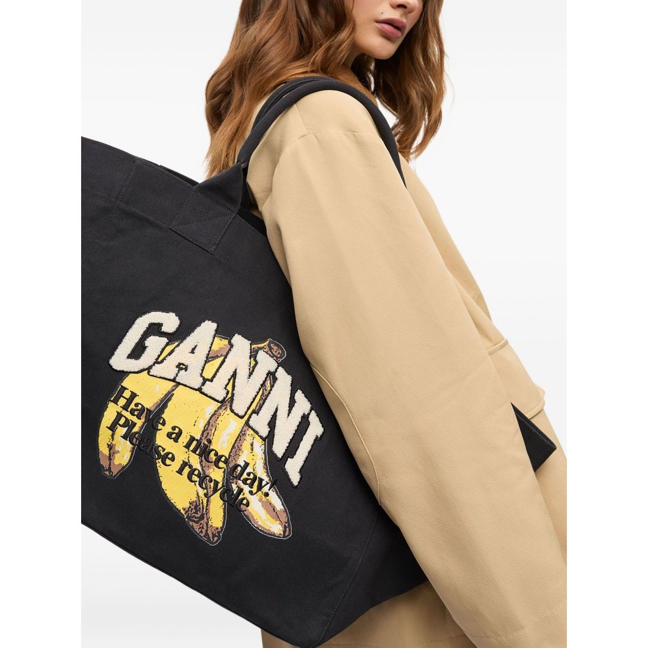 Woman wearing a beige coat holding a Ganni Organic cotton XXL tote bag with black graphic print and logo, showcasing two top handles.